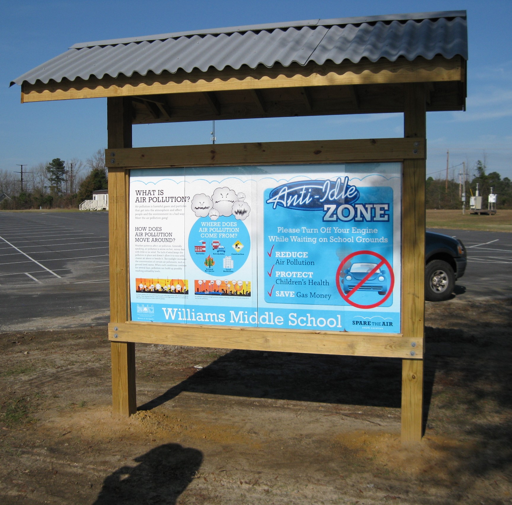 Here’s brief alt text for the image: “Outdoor wooden bulletin board displaying posters about air pollution and an anti-idling zone at Williams Middle School in a parking lot.”