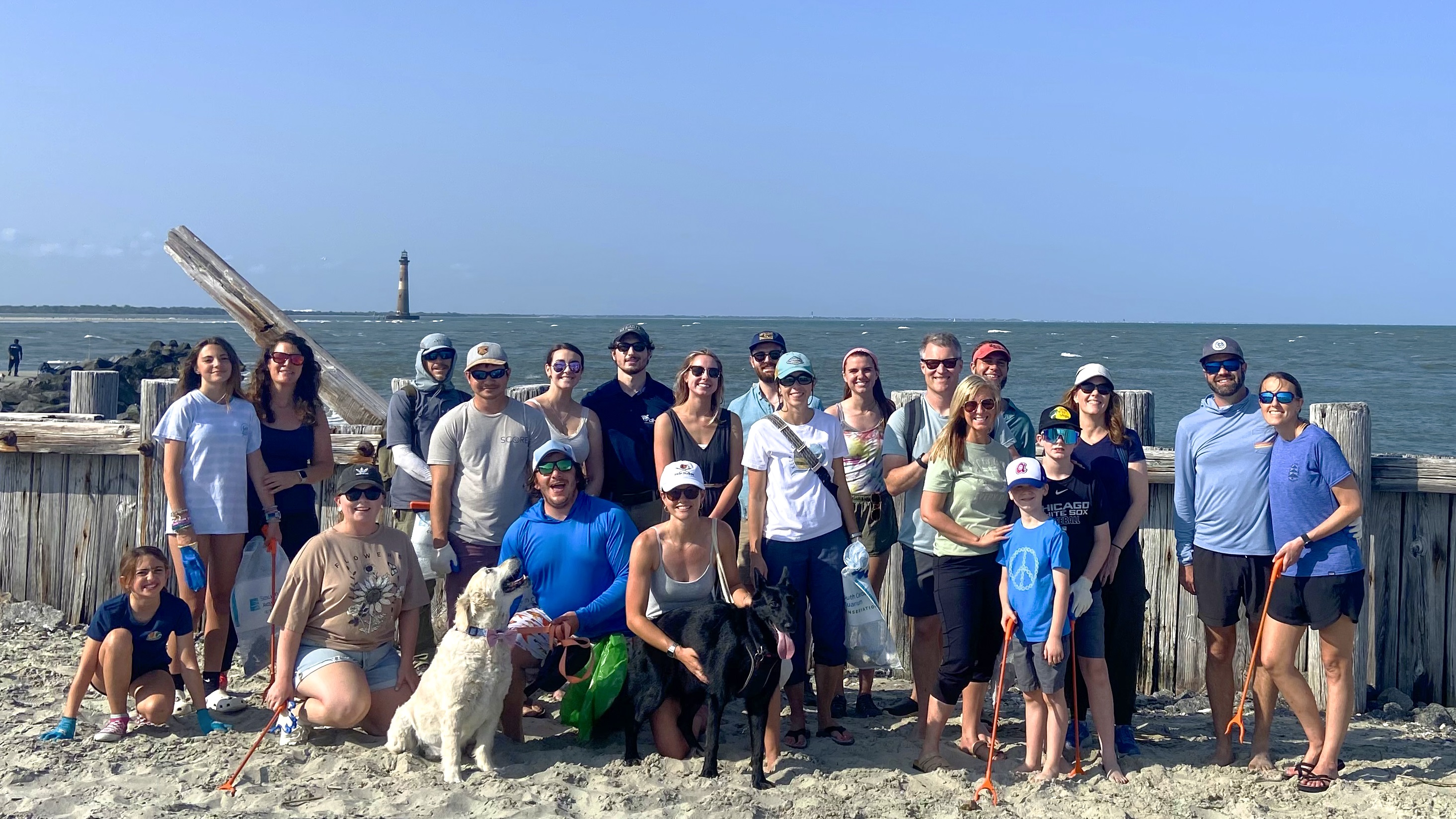 Group of people standing on a sandy beach with two dogs, holding bags and grabbers for a cleanup activity. The ocean and a distant lighthouse are visible in the background