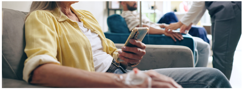 Person seated with an IV line while holding a phone, with additional individuals in treatment chairs in the background.