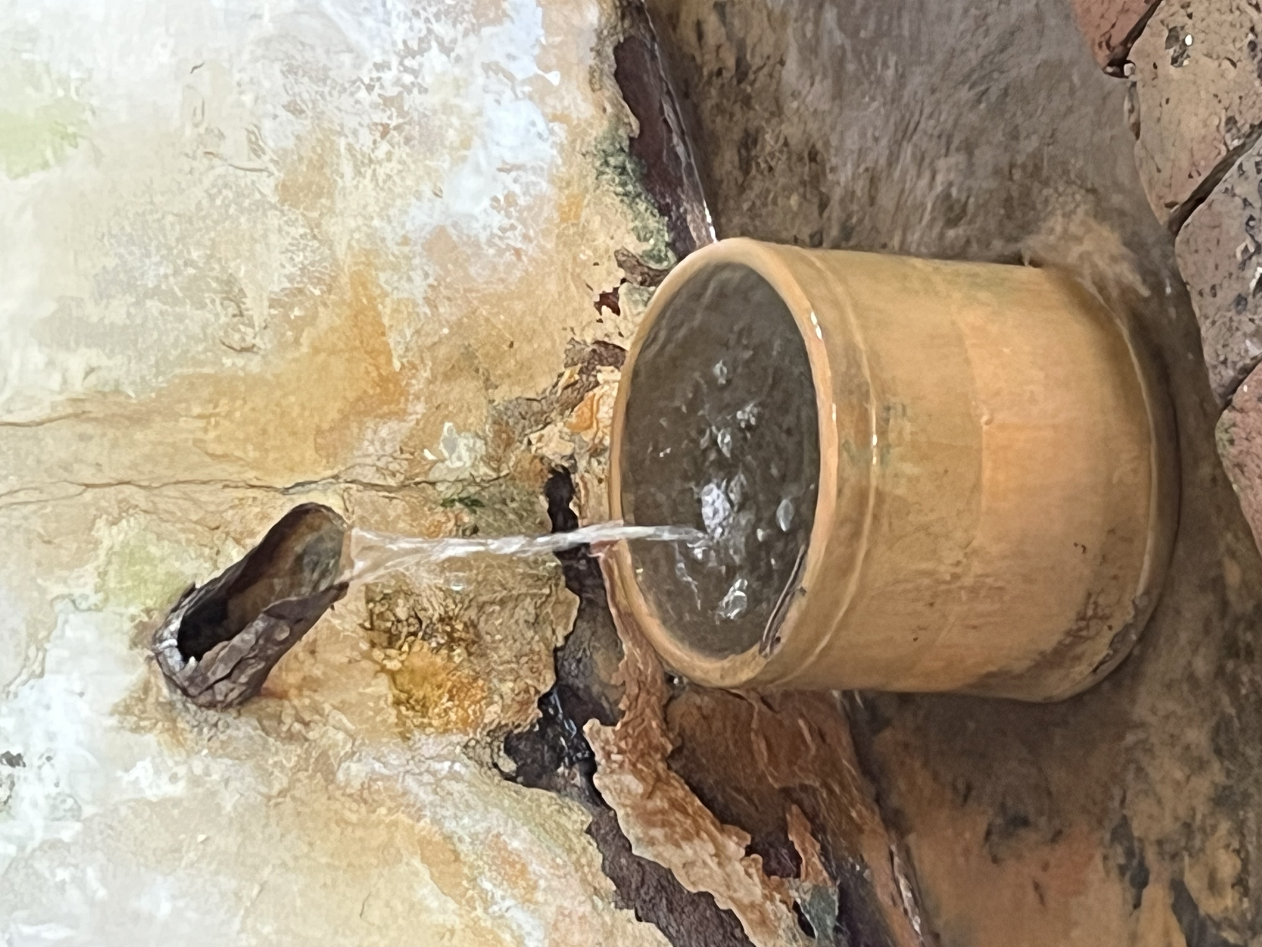 An image of a spigot with water flowing into a round basin that is sitting in some shallow water within a brick floor