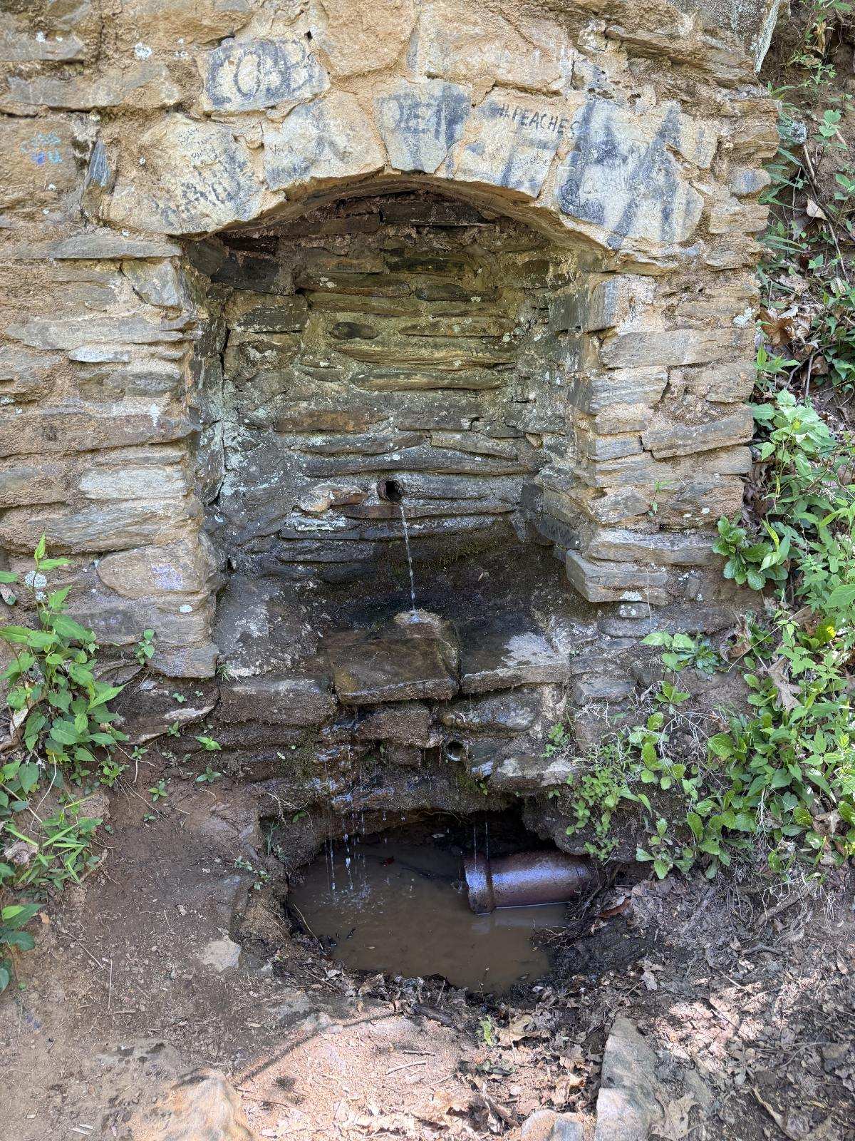 An image of Marchant Spring's stone alcove near the road with a pipe exhibiting flowing water that falls onto the ground where another pipe sits