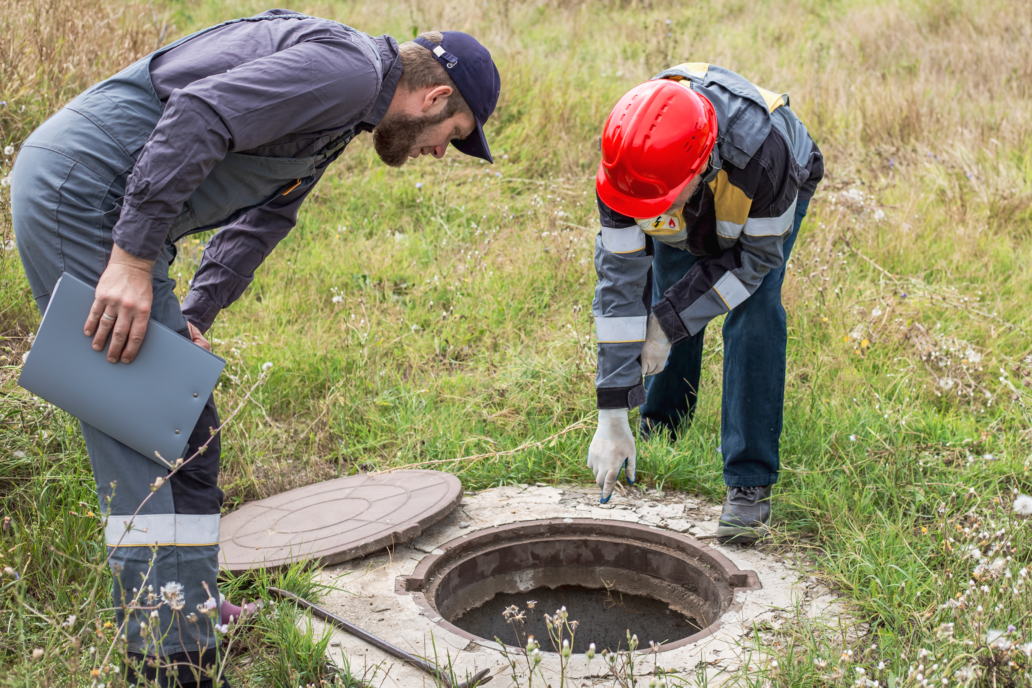 Two workers inspect an open septic tank in a grassy field.