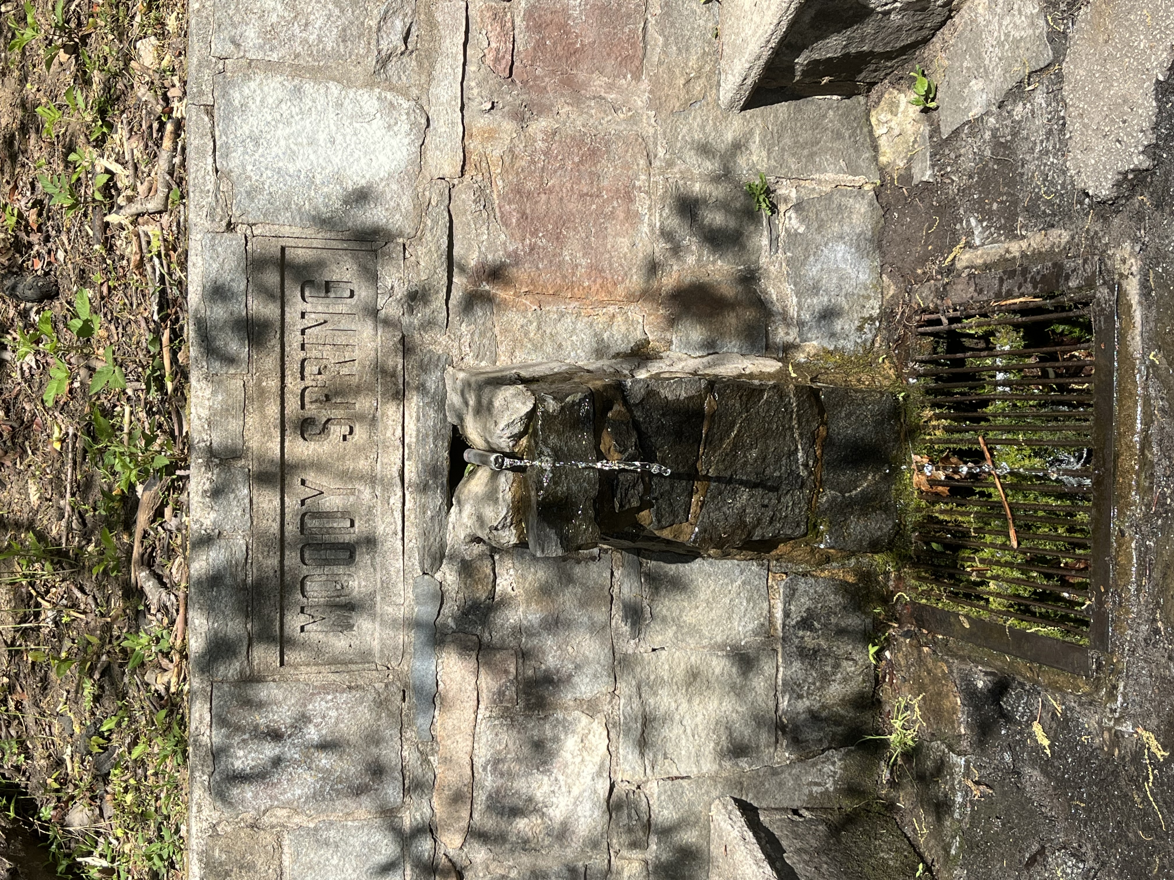 Frontal image of water flowing out of a stone structure with the words Moody Spring above the spout with a mossy grate below and stone benches on either side