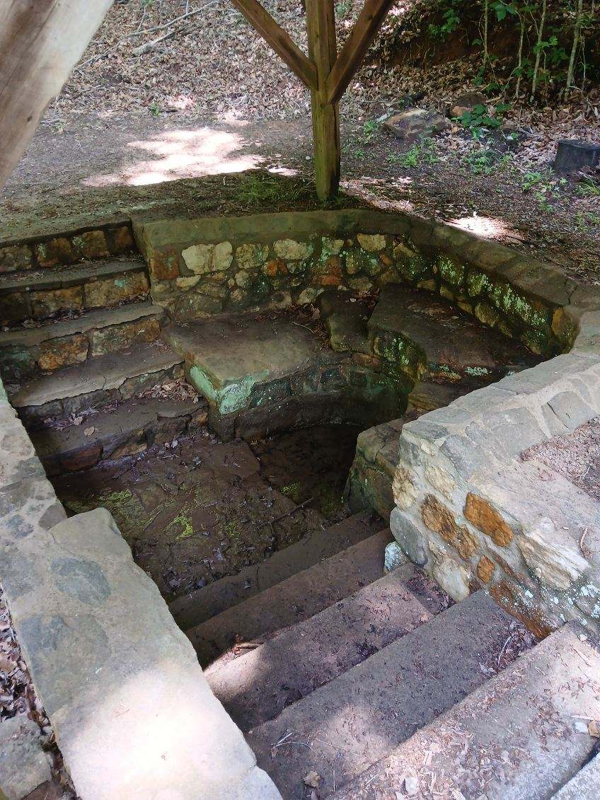 An image of a low depression surrounded by stone steps under a park shelter where water flows out of the nearby hill and continues out from underneath the structure
