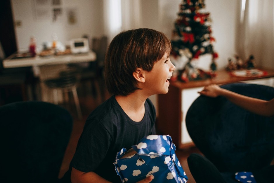 A child smiling while holding a gift wrapped in blue paper with white hearts, with a decorated Christmas tree in the background.