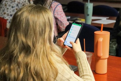 A person with long hair sits at a table, looking at a smartphone displaying a messaging app. In front of them are two tall cups, one orange and one clear. Other people are seated at tables in the background.