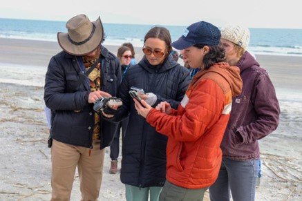 A group of people standing on a beach are examining handheld devices. The ocean is in the background. The individuals are dressed in winter clothing, including hats and jackets.