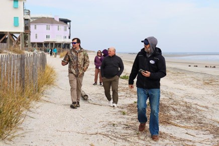 A group of people walking along a sandy beach near wooden houses on stilts. The area is bordered by a wooden fence and grassy dunes. The sky is clear, and the sea is visible in the background.