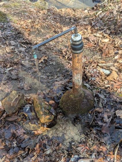 An image of a rust colored pipe with an extended smaller pipe that has water flowing out of it and the area has many leaves nearby