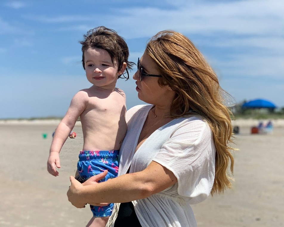 A person with long hair holds a child at a beach. The child is wearing colorful swim shorts. In the background, there are beach umbrellas and a clear blue sky.
