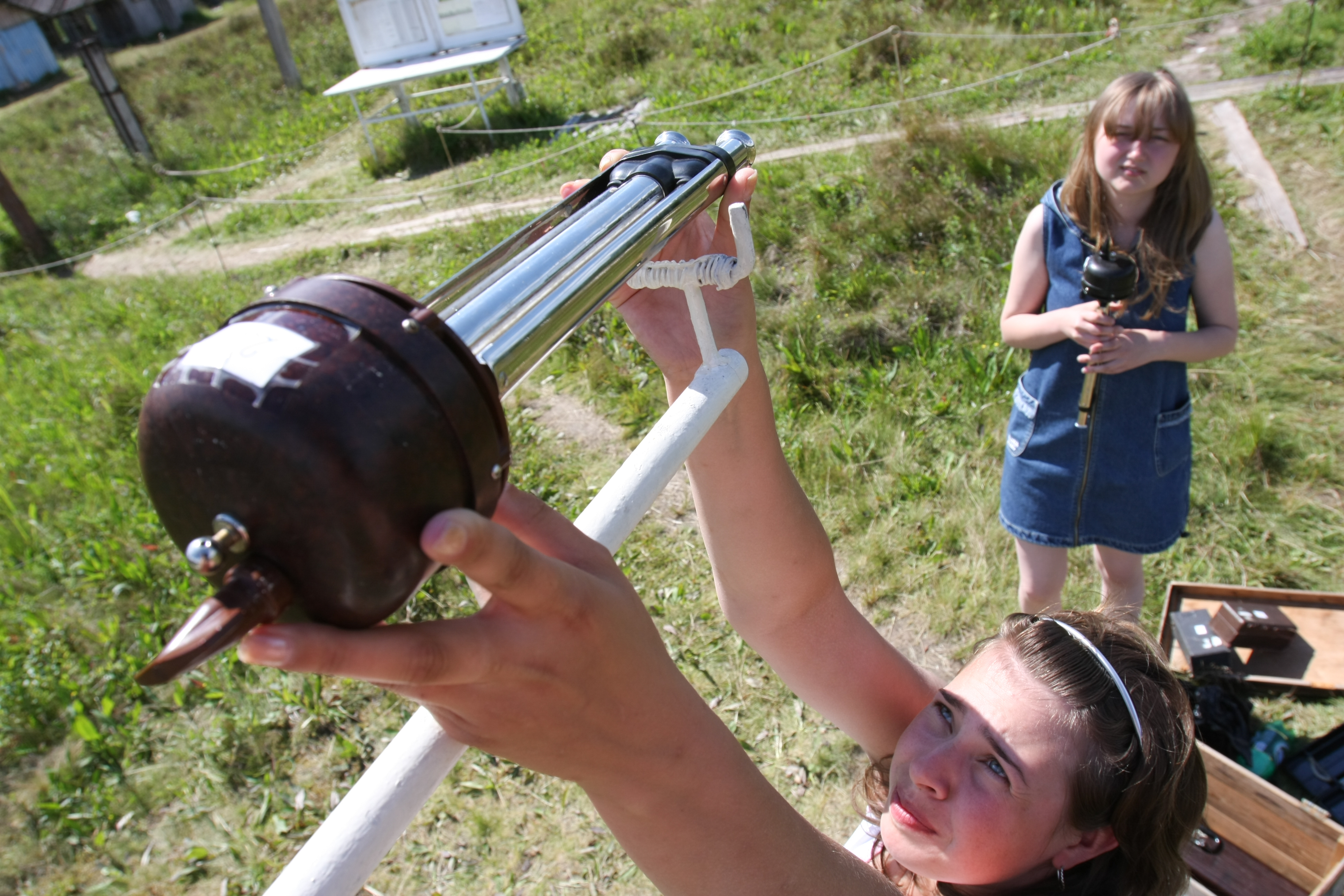 Two individuals outdoors in a grassy area are working with long metal instruments, one adjusting a mounted device while the other stands nearby holding similar equipment.