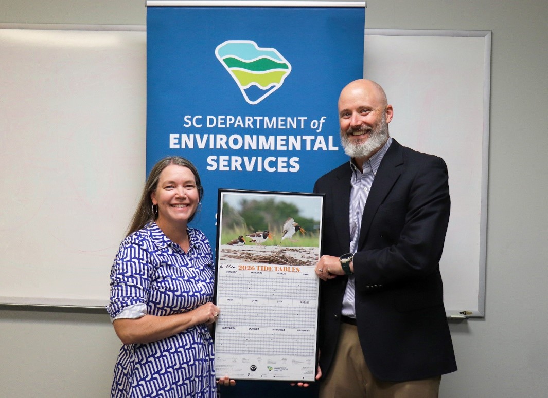 Two individuals standing in front of a banner that reads “SC Department of Environmental Services” while holding a large 2026 tide tables poster featuring a bird photograph at the top. The banner displays a stylized map graphic, and a whiteboard is visible in the background.