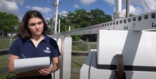 A person with short hair stands outside holding a notepad, near a large measuring instrument.