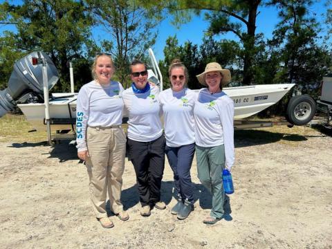 Four people in matching shirts with SCDES logos standing arm in arm in front of a boat on a sunny day.