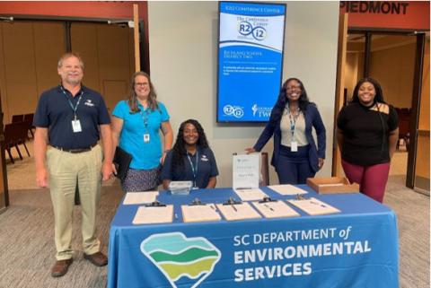 Five individuals standing behind a table with informational materials at the SC Department of Environmental Services booth in a conference center.