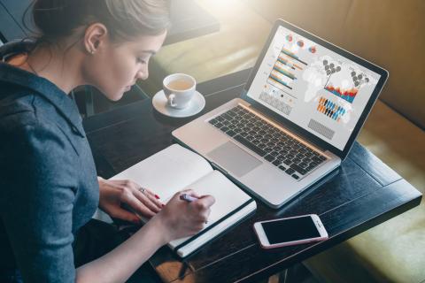 Young business woman sitting at table and taking notes in notebook. On computer screen graphics and charts. Side view. On table is laptop, smartphone and cup of coffee