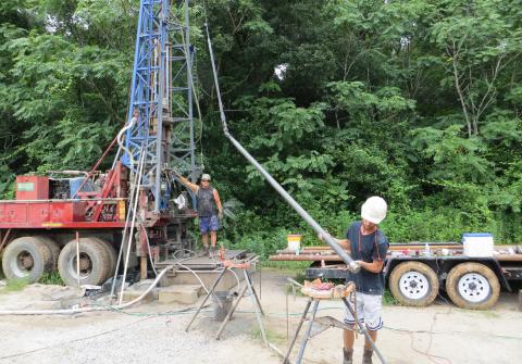 Two individuals are working at a drilling site in a forested area. One operates equipment near a large red and blue drilling rig mounted on a truck, while the other manages a long pipe.