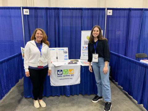 Two individuals standing in front of a booth with blue curtains at an event. A table between them displays brochures, flyers, and educational materials, with a banner on the front that reads “take action SC” and shows recycling‑related icons.