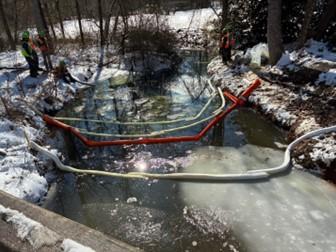 Workers in safety gear stand near a partially frozen stream where containment booms have been placed in the water. Snow covers the ground and surrounding trees, and the booms form barriers across the icy surface to control a spill