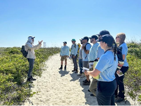 A group of people standing on a sandy trail surrounded by low vegetation while listening to another person who is speaking and gesturing with both hands. The group is outdoors under a clear blue sky, and some individuals are holding notebooks or papers.